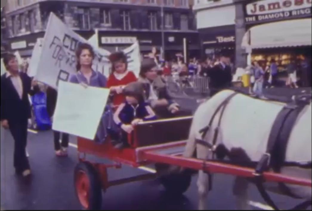 A still image from a video of a protest march in Dublin. In the foreground, Nan Joyce and her children are sitting in a horse-led cart. She's holding a sign (not legible in the image). Other marchers walk behind her with a large banner (partially legible: Commitee For...).