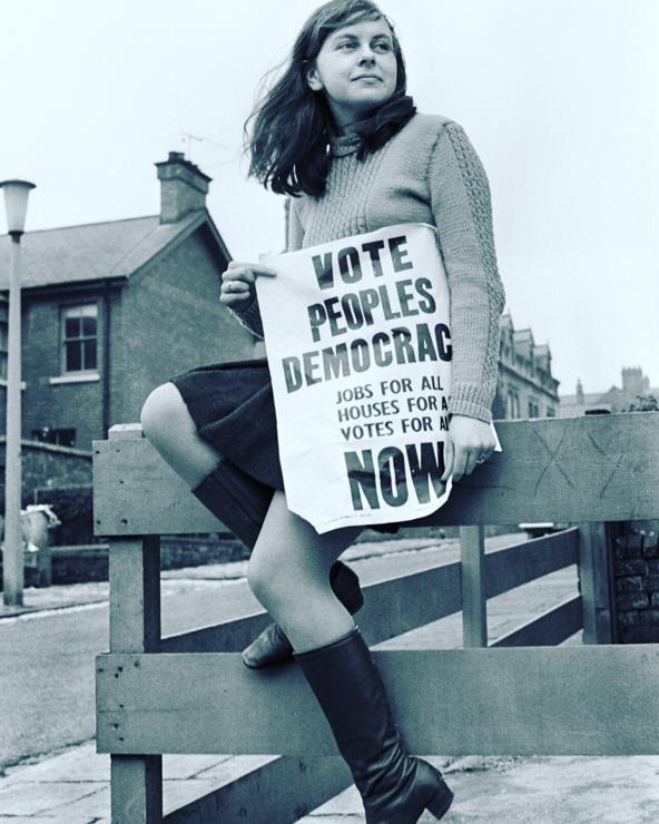 Young woman sits on a fence in a residential area holding a sign that says Vote Peoples Democrac
Jobs for All
Houses for All
Votes for All
NOW