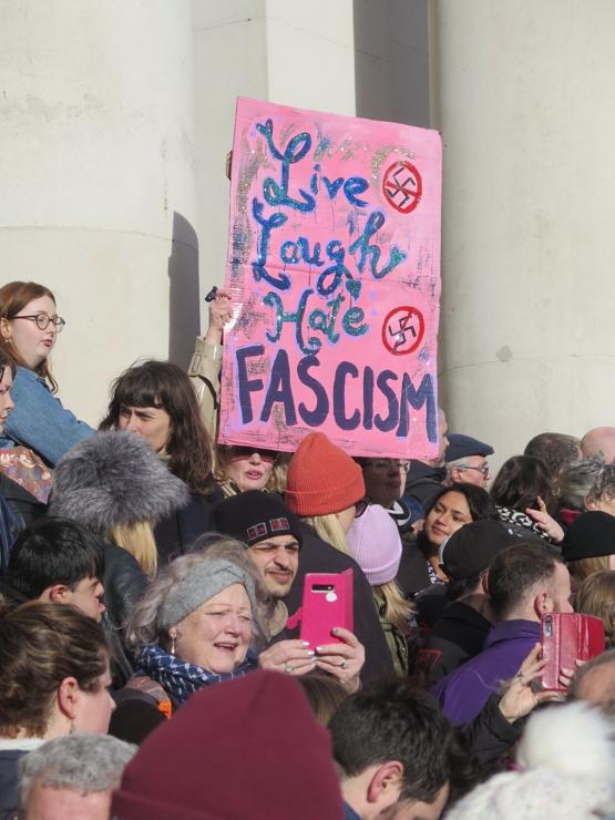 People at a demonstration. A woman holds up a home-made sign saying:

Live
Laugh
Hate
FASCISM

with pictures of crossed-out swastikas.

In front of her another woman is taking a picture with a smartphone.