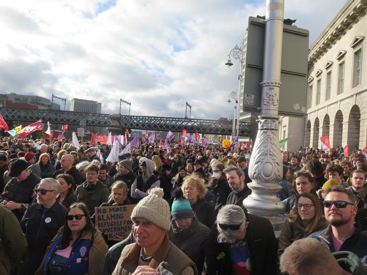 Large number of people at a demonstration with various flags flying though the symbols and text on them is not so clear. In the foreground a woman holds a hand drawn sign saying "No more blaming refugees for Government fuck ups". 

Behind the demonstration we can see the Loop railway bridge across the Liffey, while to the right is an ornate lamp-post and the ornate neoclassical frontage of the Custom House.