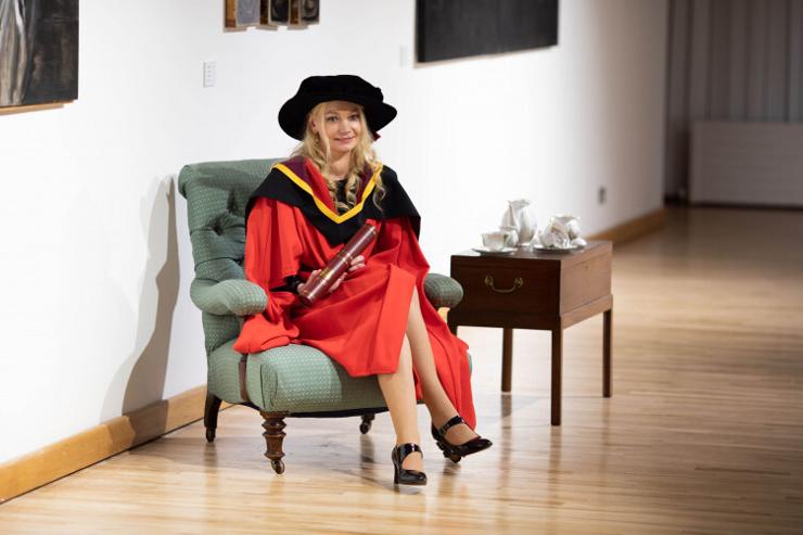 A photograph of Dr Sindy Jouce, lecturer in sociology at the University of Limerick. She is seen in a white-walled room sat in a pale green chair. She is wearing red, black and yellow formal graduation gowns and is holding a wooden cylinder upon her doctorate graduation
